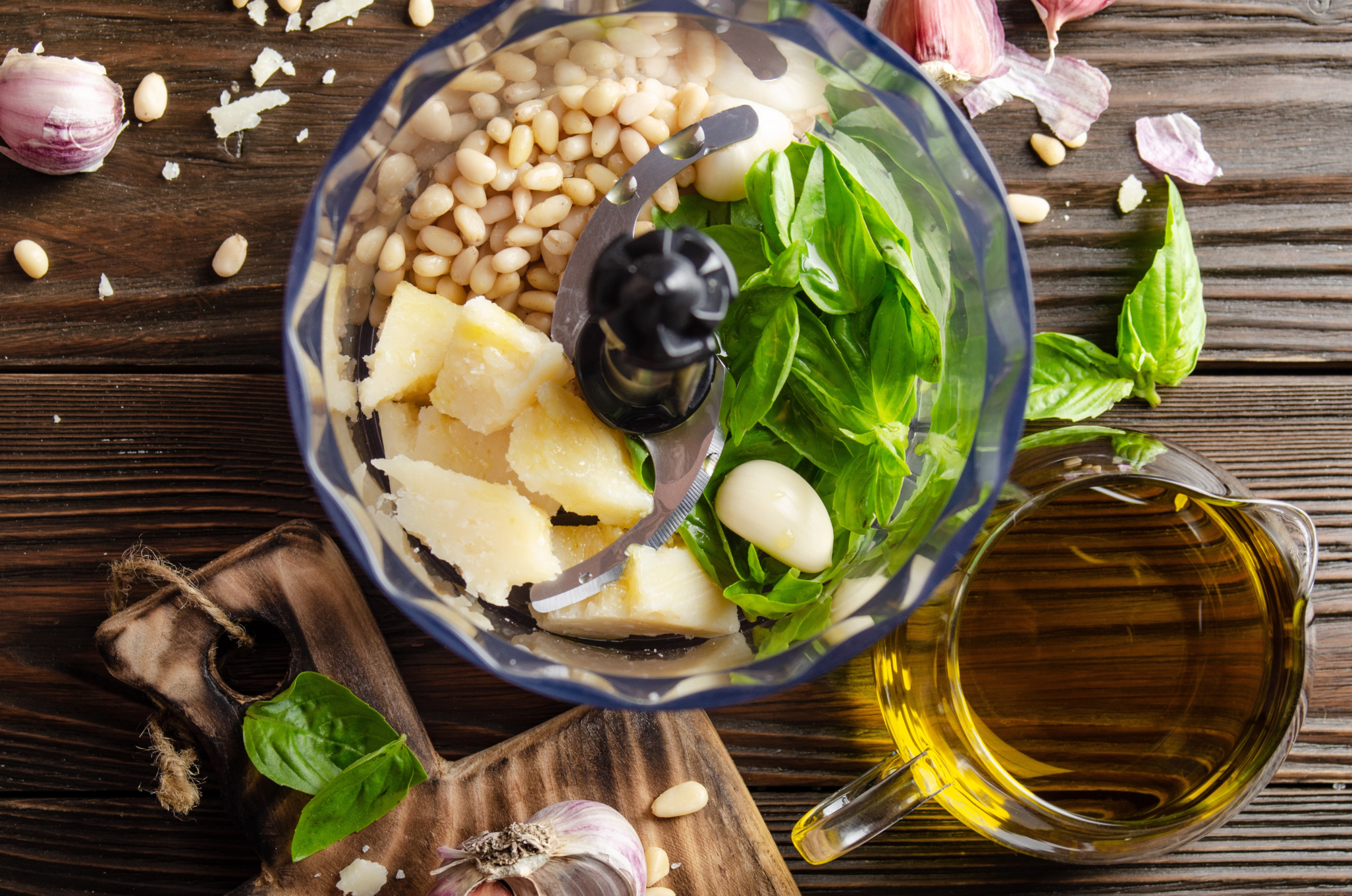 food processor with unprocessed ingredients for pesto including basil garlic pine nuts and Parmesan cheese