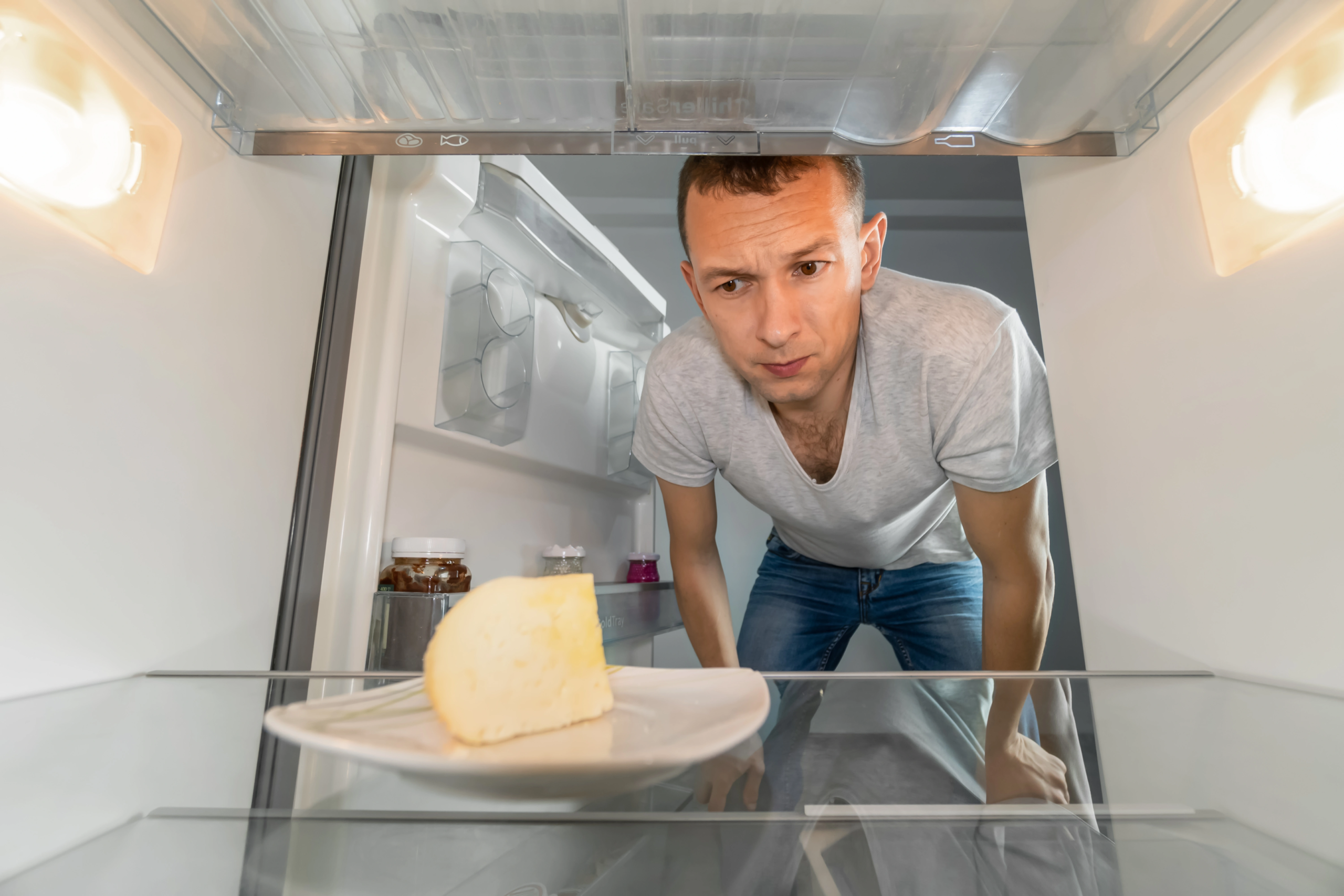 man looking inside empty fridge at a plate with only a block of cheese