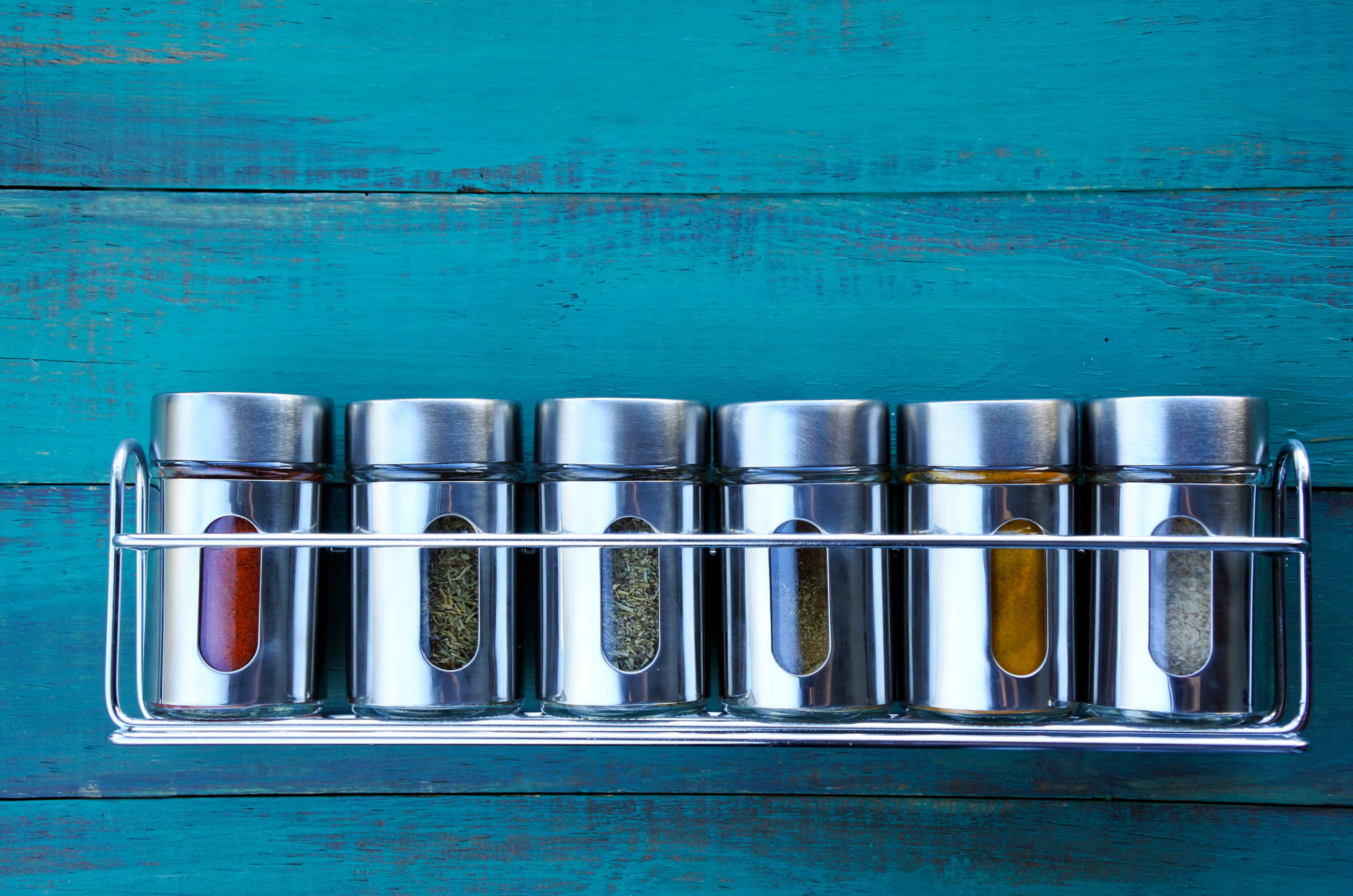 metal wall anchored spice rack with six metal canisters of spices on blue wooden background.